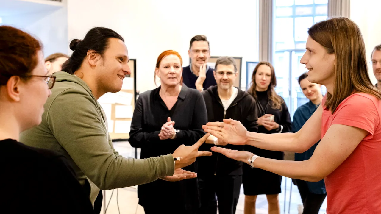 Zwei Teammitglieder spielen Schere-Stein-Papier, umgeben von applaudierenden Kolleg:innen – Symbol für gemeinsames Entscheiden und Teamstärkung
